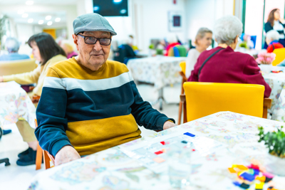 elderly man wearing glasses and a cap sitting at a table in a bright communal area surrounded by other seniors engaging in activities showcasing companionship and community spirit 4 wellness activities